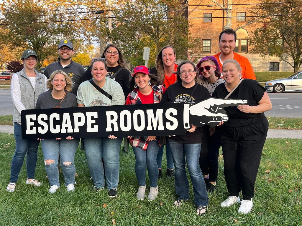 group photo of 11 people holding up an escape room sign on the yard of crooked cottage escape rooms