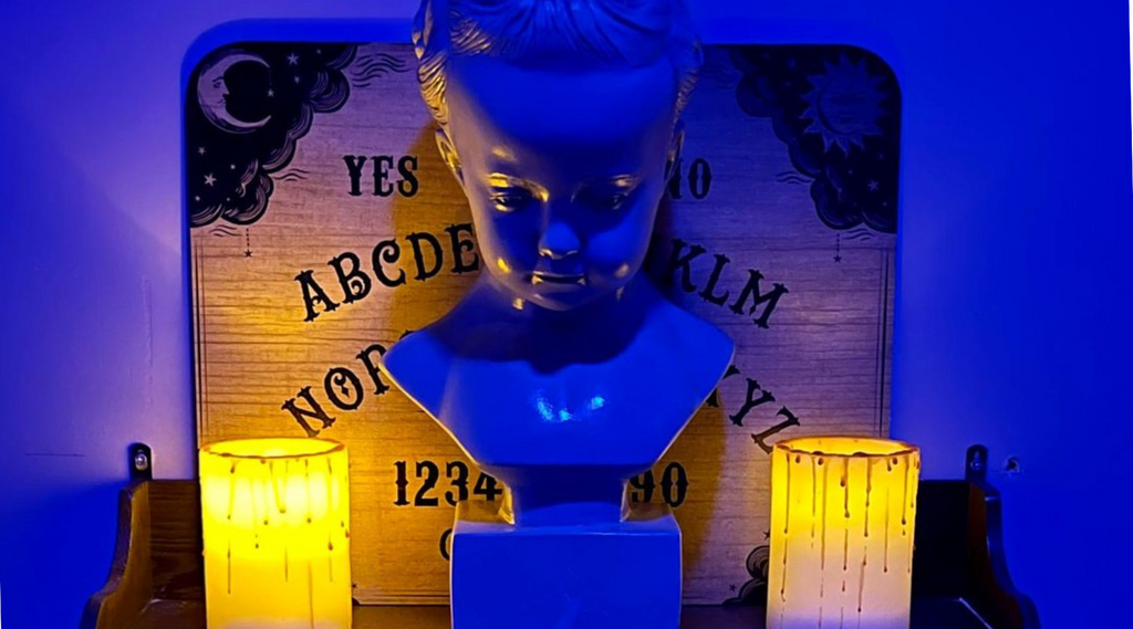 bust of a little girl sitting on a shelf between two dripping lit candles and a ouija board sitting in the background